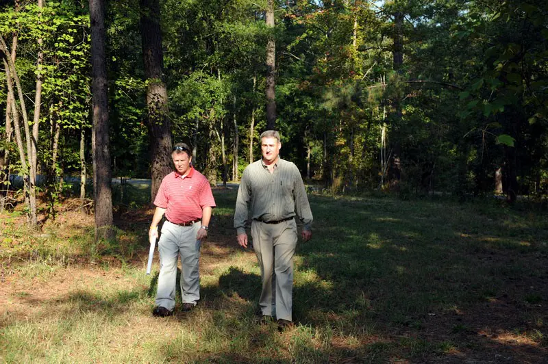 Two men standing in a wooded area, one in a red polo and the other in a gray shirt, both looking at the camera.