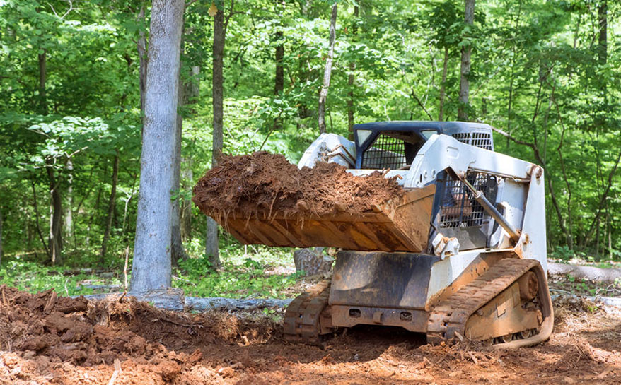 A skid steer loader carrying a load of dirt in a forested area.