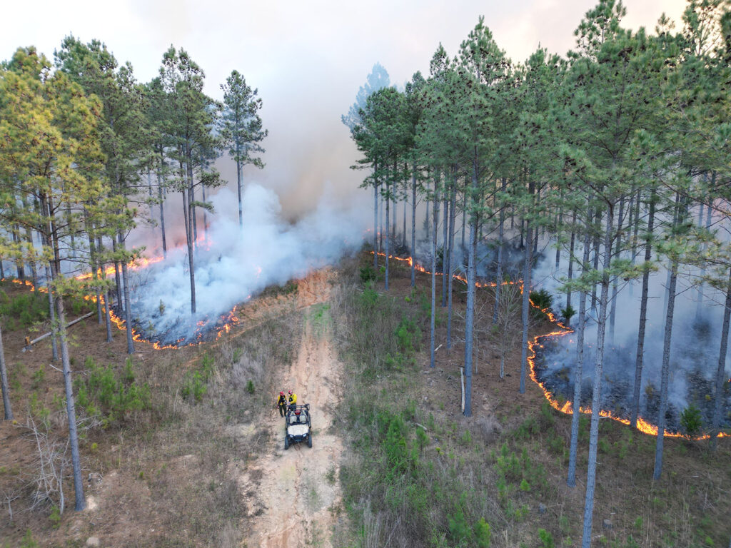 Aerial view of a forest fire with firefighters in an atv on a dirt road, smoke rising and flames spreading through trees.