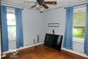 Cozy room featuring a ceiling fan, blue curtains, a TV on the floor, and light blue walls with some wear. Cozy room featuring a ceiling fan, blue curtains, a TV on the floor, and light blue walls with some wear.
