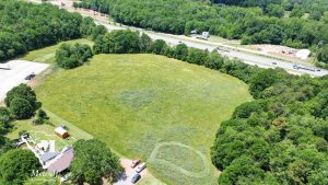 Aerial view of a multi-use property with open land adjacent to Interstate 85, surrounded by trees and parking area. Aerial view of a multi-use property with open land adjacent to Interstate 85, surrounded by trees and parking area.