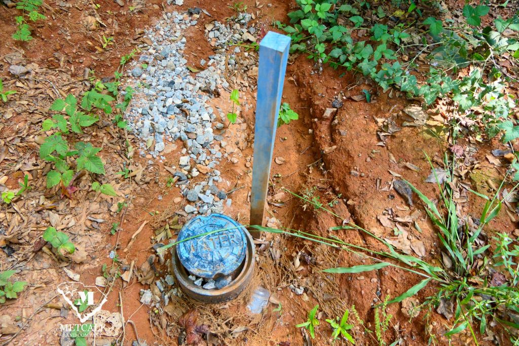Water meter and marker on a 2.5-acre homesite in Campobello, surrounded by natural vegetation and gravel.